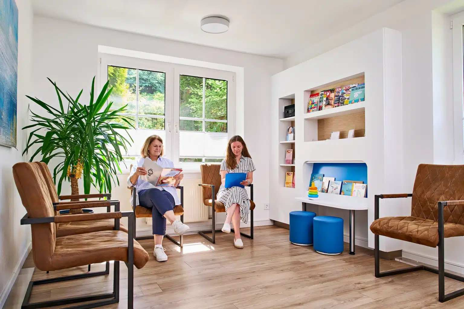 Ein heller Warteraum, in dem zwei Patient*innen – eine jüngere und eine etwas ältere – auf bequemen, gepolsterten Stühlen auf ihren Termin warten.